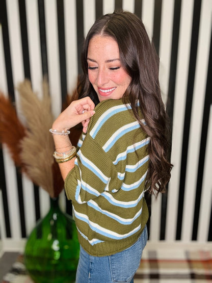A woman with long dark brown hair is shown from the back wearing an olive green knit sweater with horizontal light blue and white stripes, paired with blue jeans. She is standing in front of a black and white striped wall with a green glass vase and pampas grass in the background.