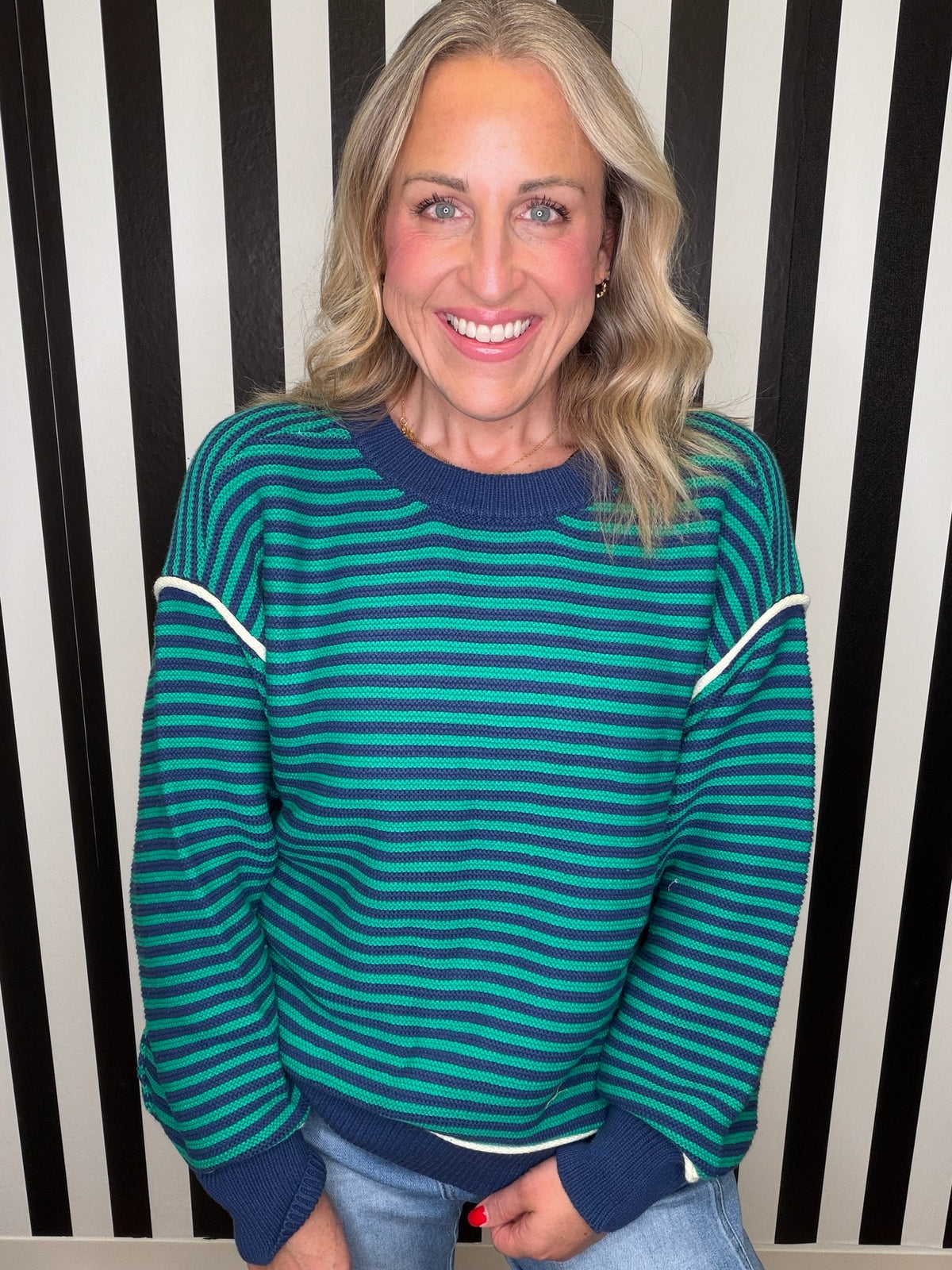 A smiling blonde woman wearing a teal and dark green striped sweater with navy blue ribbed collar, cuffs, and hem, paired with light blue jeans, standing in front of a black and white vertically striped wall.