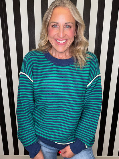 A smiling blonde woman wearing a teal and dark green striped sweater with navy blue ribbed collar, cuffs, and hem, paired with light blue jeans, standing in front of a black and white vertically striped wall.