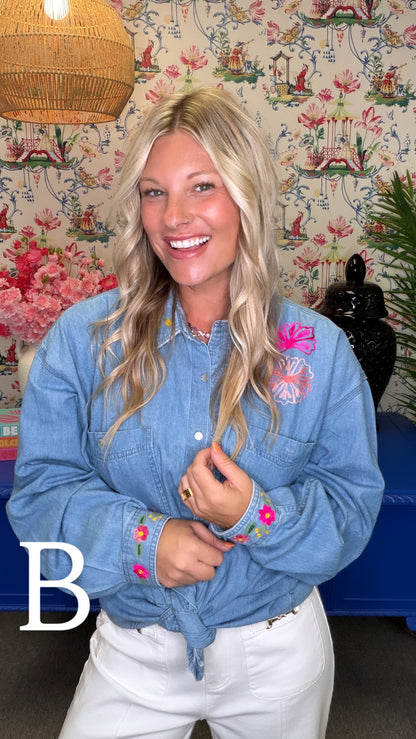 A woman with blonde hair wearing a light blue denim shirt with pink floral embroidery on the chest and cuffs, paired with white pants. She is standing in front of a floral wallpaper background with a wicker lamp visible at the top left.