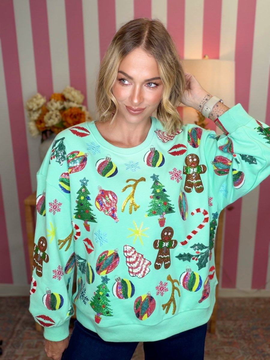 A woman wearing a mint green Christmas crewneck sweatshirt covered in colorful holiday prints including gingerbread men, ornaments, candy canes, and trees, posed in front of a pink and white striped wall.