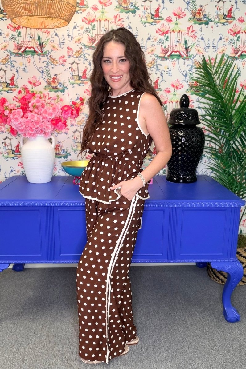 A woman modeling a brown and white polka dot sleeveless maxi dress with white piping, standing in front of a blue cabinet and floral wallpaper.