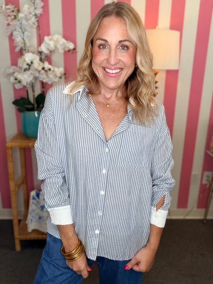 A smiling blonde woman wearing a blue and white vertically striped button-down shirt with white cuffs, paired with blue jeans, standing in front of a pink and white striped wall.