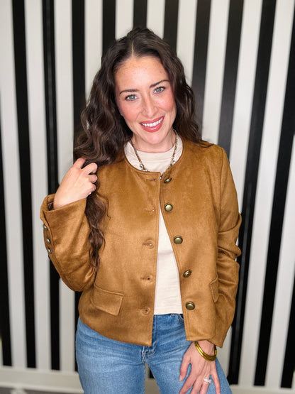 A woman with long wavy brown hair smiling, wearing a cropped tan suede jacket with gold-tone buttons over a cream turtleneck and blue jeans, standing in front of a black and white vertically striped background.