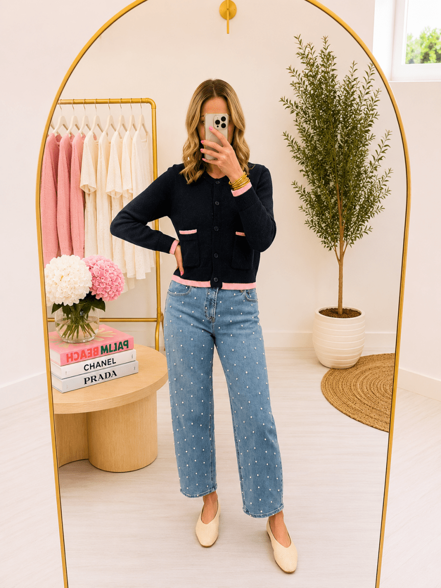 A woman takes a mirror selfie in a boutique showing a navy cardigan with pink trim and light blue polka dot jeans.