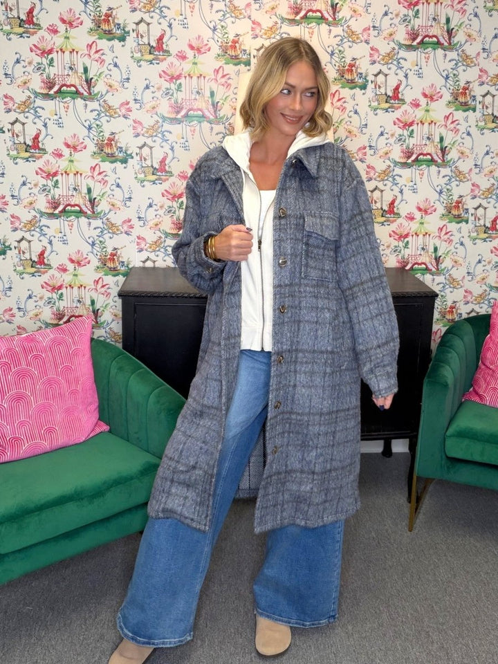A woman stands in a room with floral wallpaper, green velvet chairs, and a black cabinet, wearing a long gray plaid button-up coat over a white top and wide-leg blue jeans.