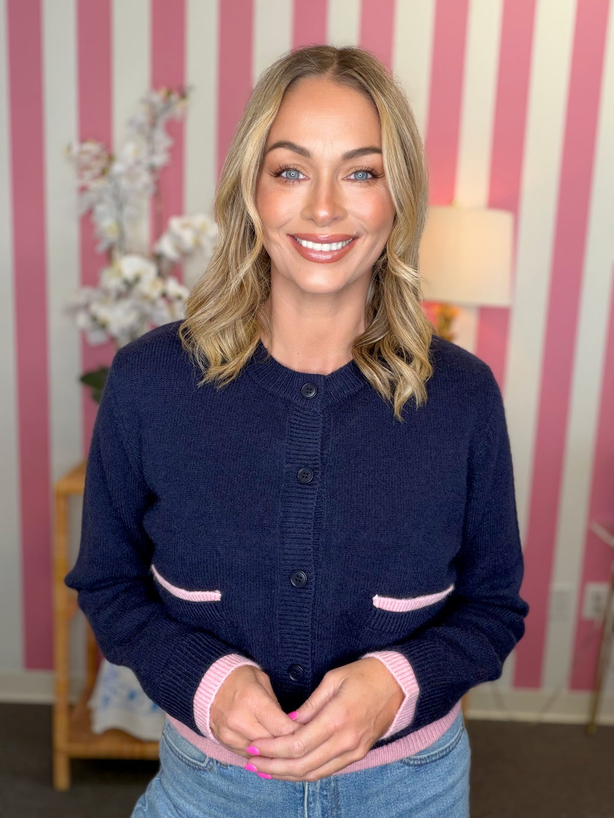 A smiling blonde woman wearing a navy blue knit cardigan with light pink trim on the pockets, cuffs, and hem, paired with blue jeans. She is standing in front of a pink and white striped wall.
