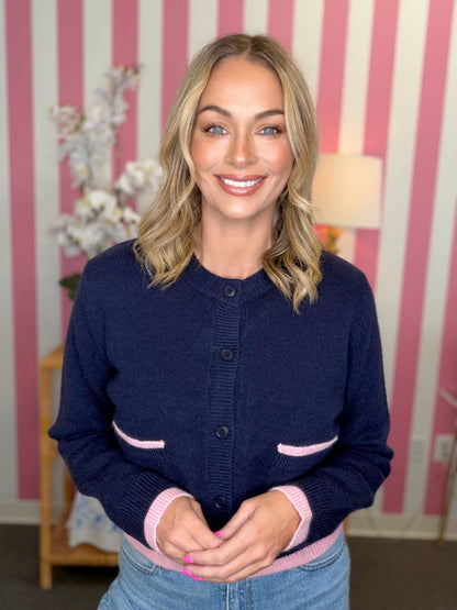 A smiling blonde woman wearing a navy blue knit cardigan with light pink trim on the pockets, cuffs, and hem, paired with blue jeans. She is standing in front of a pink and white striped wall.