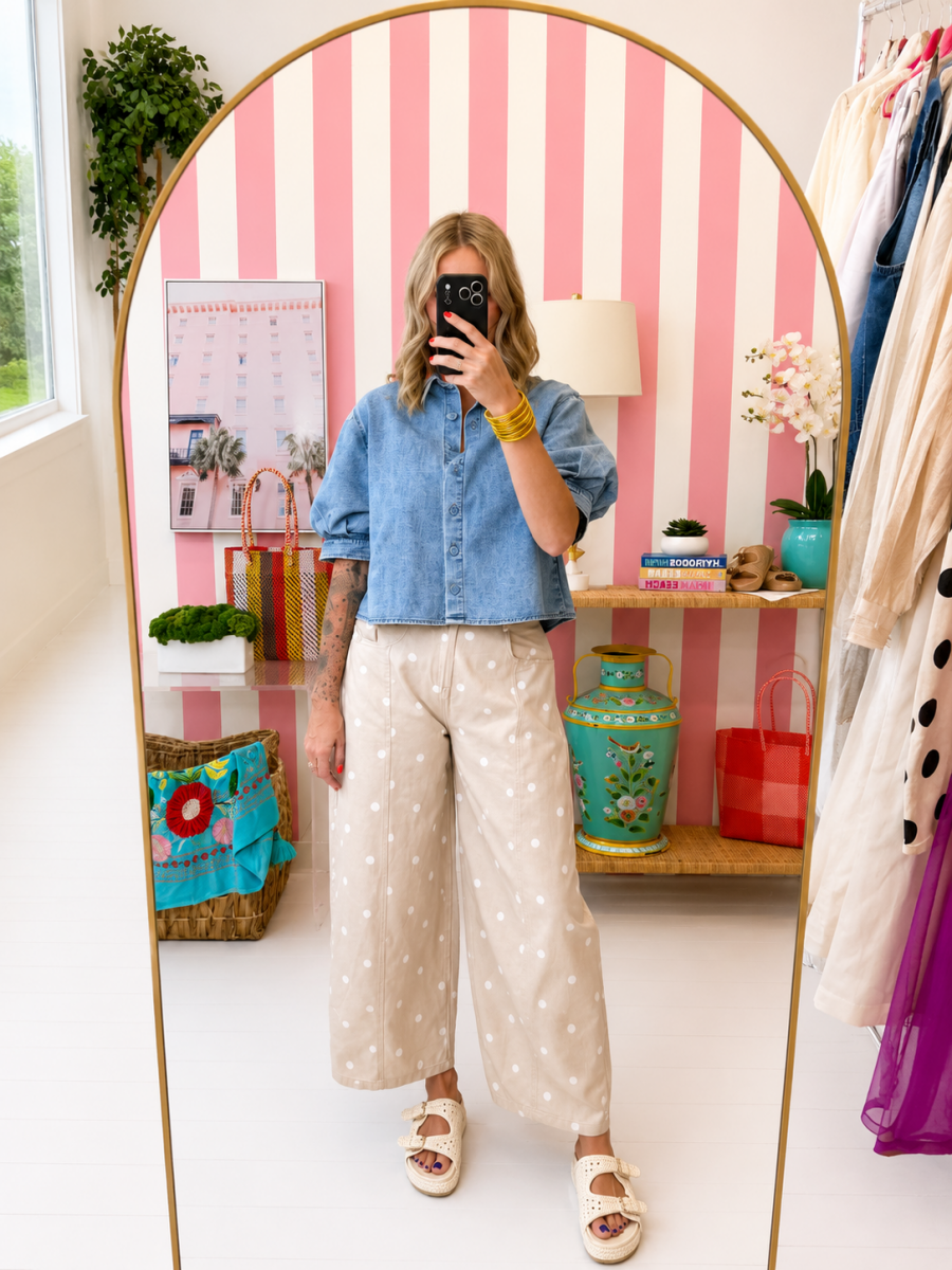 Person taking a mirror selfie wearing a denim shirt and polka dot pants in a room with striped walls and shelves.