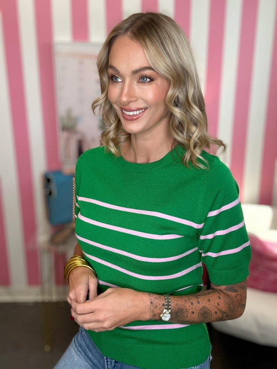 A woman with blonde wavy hair wearing a green short-sleeve knit sweater with white horizontal stripes, paired with blue jeans. She is standing in front of a pink and white striped wall.