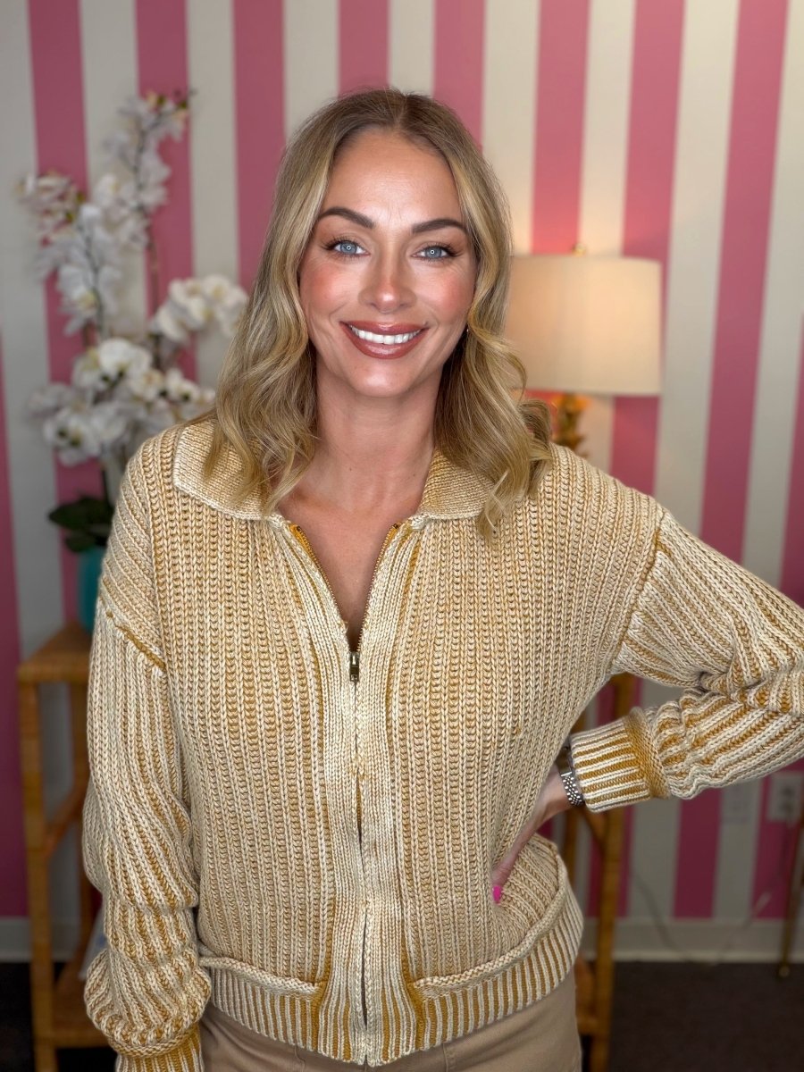 A smiling blonde woman wearing a textured zip-up knit cardigan in a warm cream and mustard yellow color, standing in front of a pink and white striped wall.