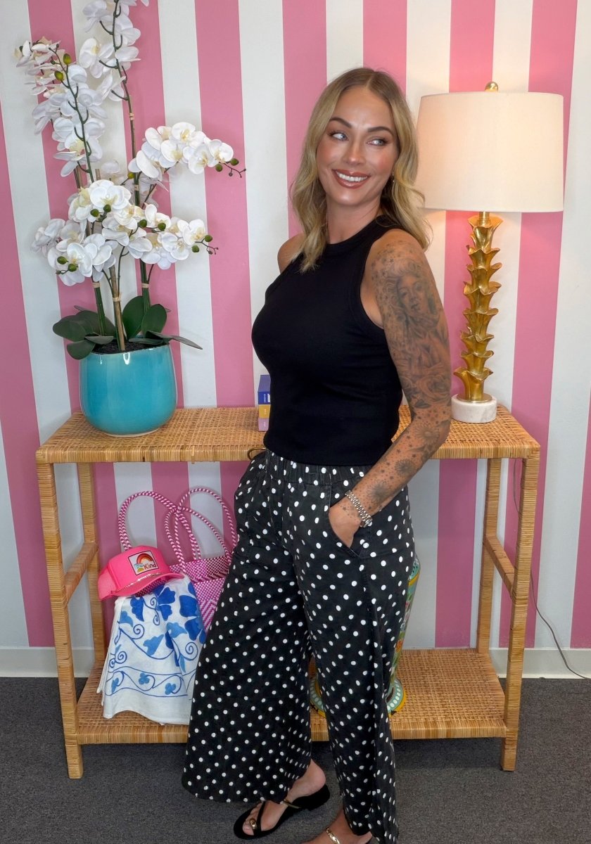 A woman posing in a black sleeveless top and black pants with white polka dots, standing in front of a pink and white striped wall next to a wooden console table with a lamp and a turquoise pot of white flowers.