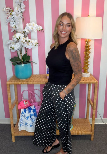 A woman posing in a black sleeveless top and black pants with white polka dots, standing in front of a pink and white striped wall next to a wooden console table with a lamp and a turquoise pot of white flowers.