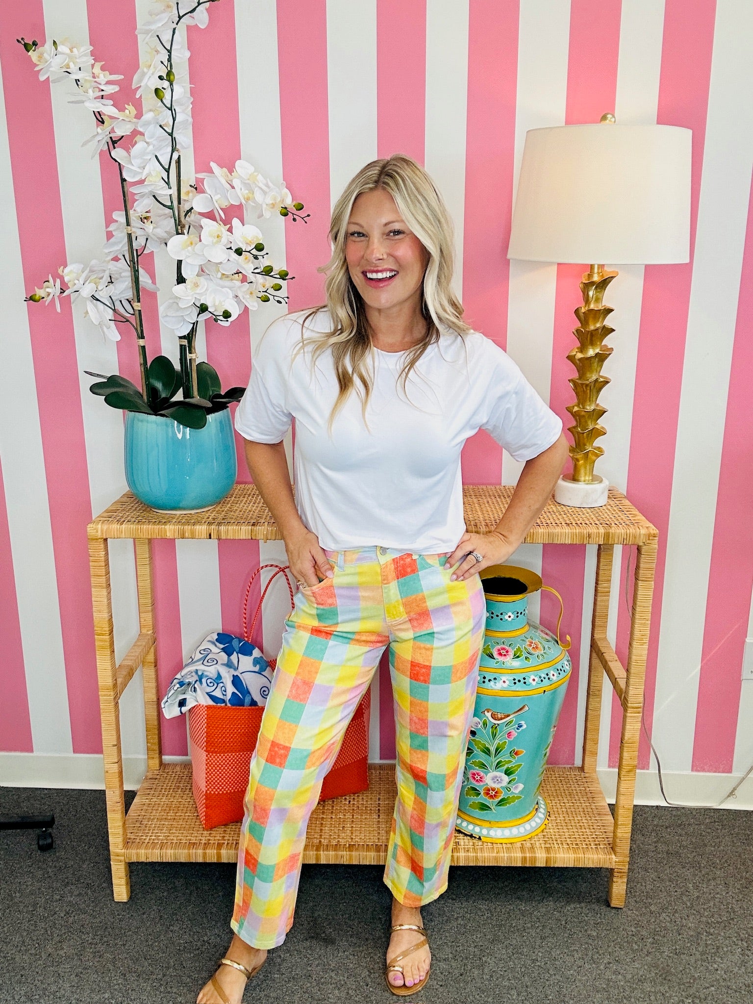 Woman in a white t-shirt and colorful pants standing in front of a pink and white striped wall with a table and decorative items.