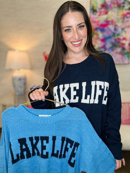 Woman holding a blue 'Lake Life' sweater in a room with colorful decor.