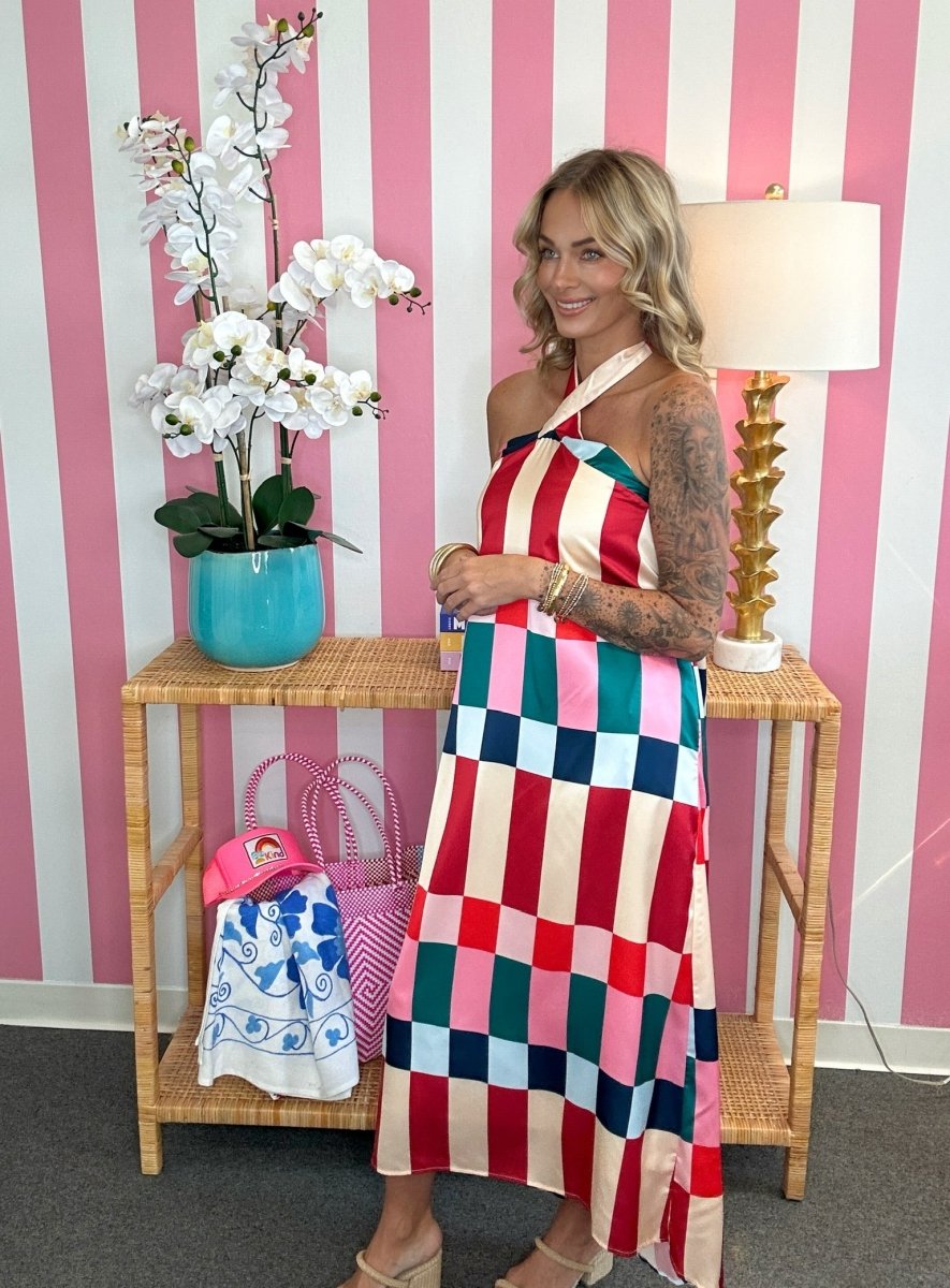 A woman models a colorful halter-neck maxi dress with a large checkered pattern in pink, red, blue, green, and cream, standing in front of a pink and white striped wall next to a side table with a lamp and flowers.