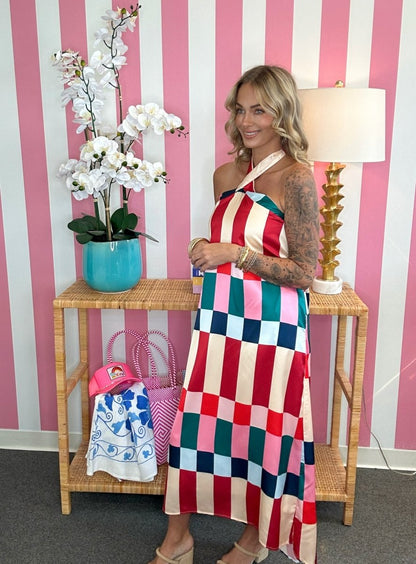 A woman models a colorful halter-neck maxi dress with a large checkered pattern in pink, red, blue, green, and cream, standing in front of a pink and white striped wall next to a side table with a lamp and flowers.