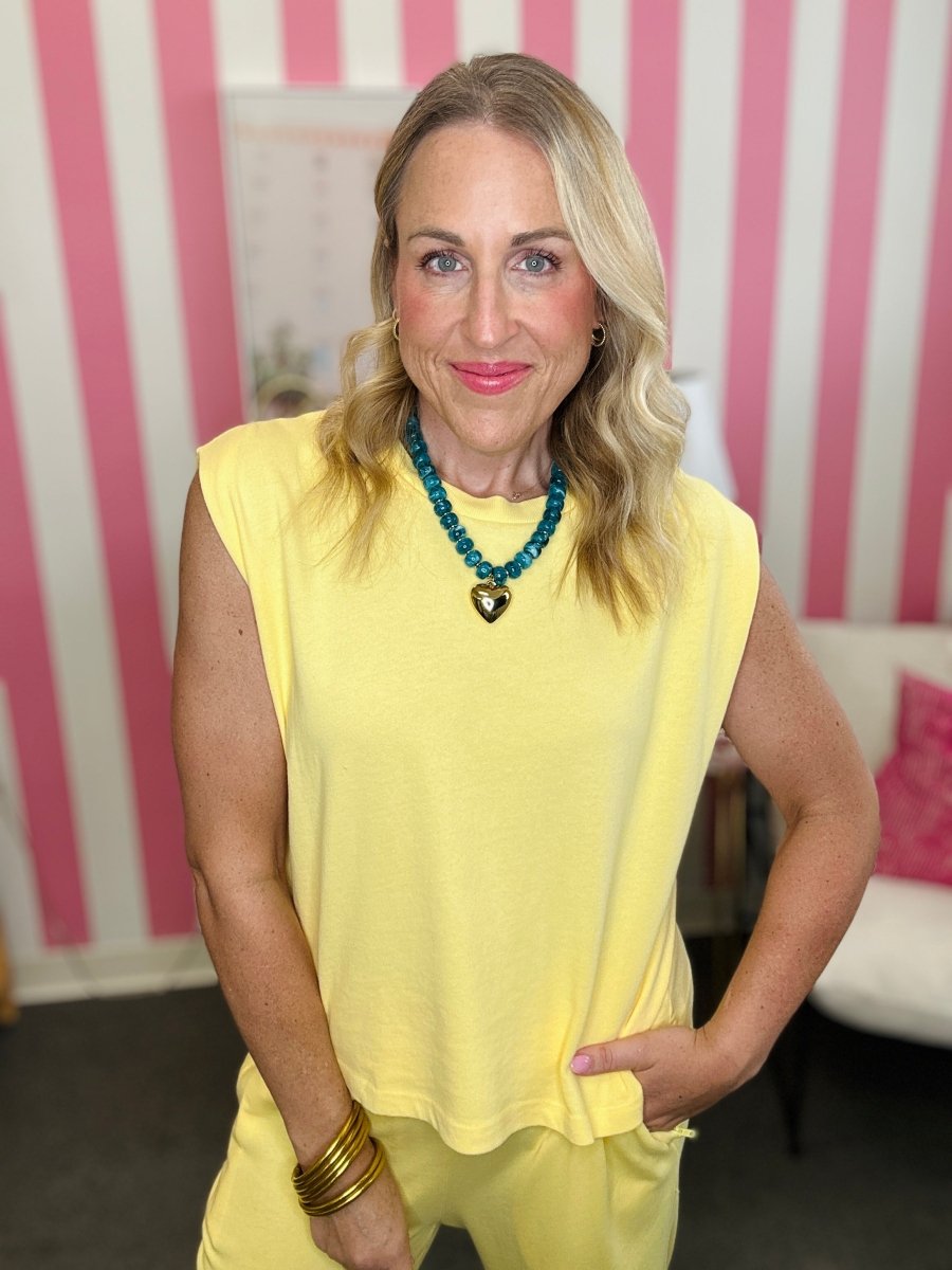 A woman with blonde hair wearing a pale yellow sleeveless top and matching yellow pants, accessorized with a turquoise beaded necklace and gold bracelets, standing in front of a pink and white striped wall.