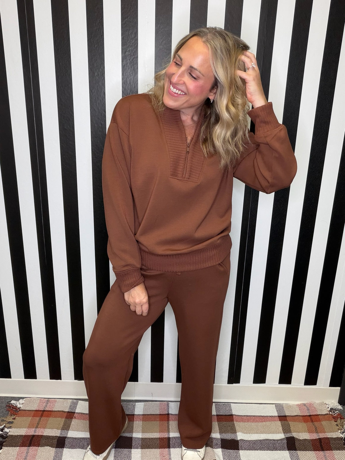 A woman modeling a matching brown knit lounge set consisting of a long-sleeve half-placket top and wide-leg pants, standing in front of a black and white striped wall.