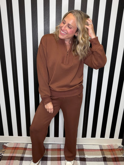 A woman modeling a matching brown knit lounge set consisting of a long-sleeve half-placket top and wide-leg pants, standing in front of a black and white striped wall.