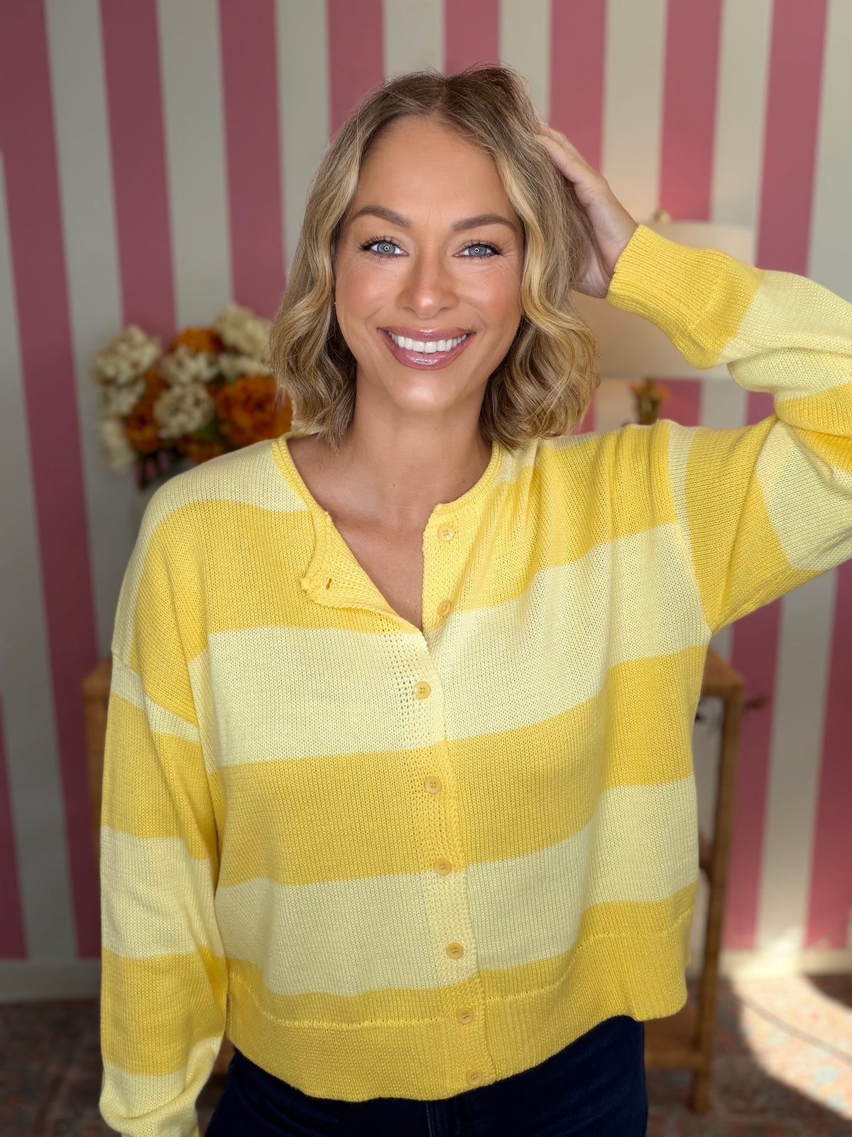 A smiling blonde woman wearing a yellow and cream striped knit cardigan sweater, posing in front of a pink and white striped wall.
