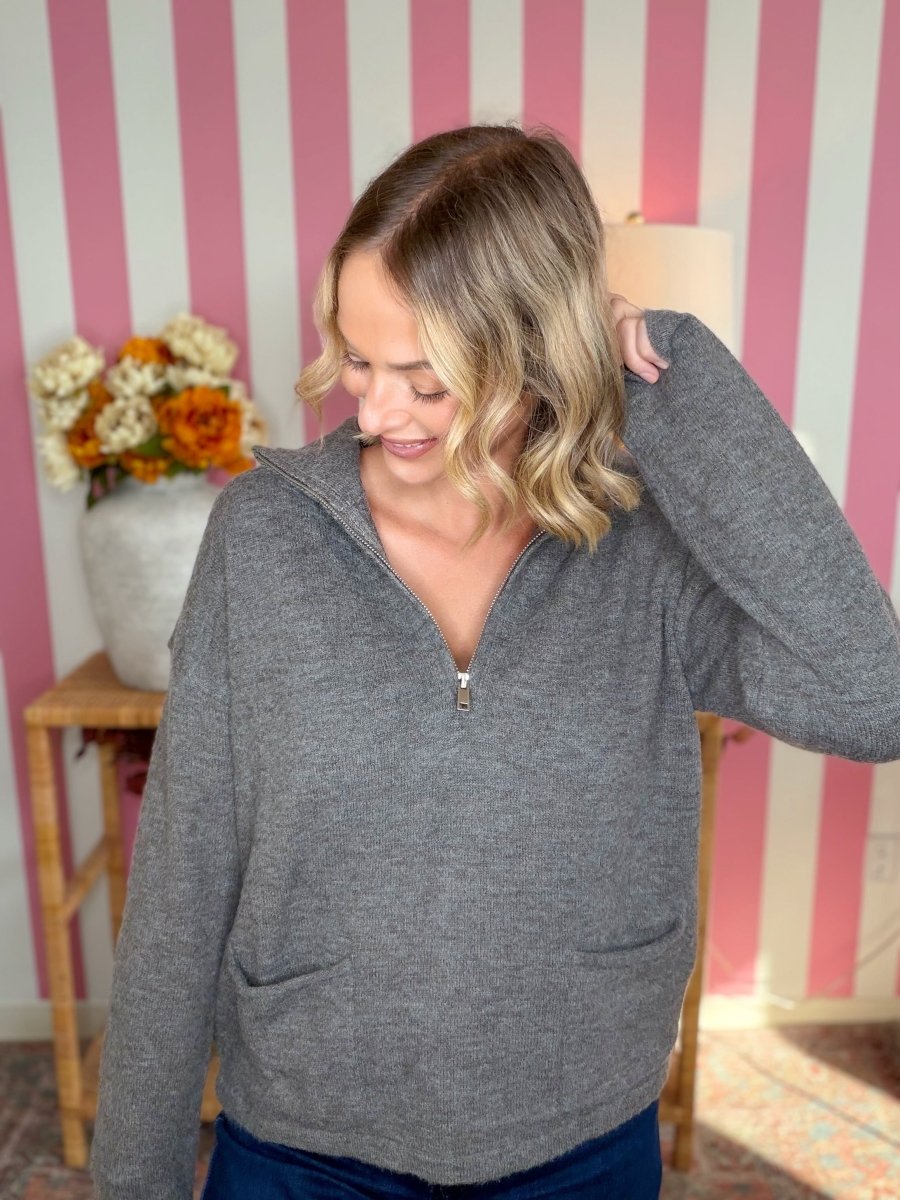 A woman wearing a heather gray half-zip hoodie with a split neckline and front pockets, standing in front of a pink and white striped wall.