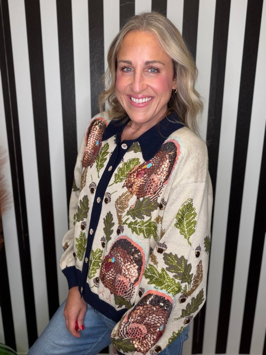 A smiling woman with blonde hair wearing a cream-colored button-up cardigan with a navy blue collar and cuffs, decorated with sequined woodland motifs including acorns, leaves, and mushrooms, paired with blue jeans against a black and white striped background.