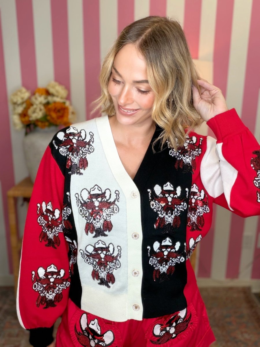 A woman wearing a red, black, and white cardigan with cowboy illustrations, posed in front of a pink and white striped wall.