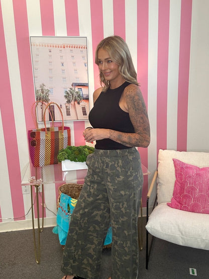 Woman in black top and camouflage pants standing in a room with pink and white striped walls.