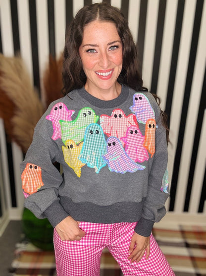 A woman wearing a gray crewneck sweatshirt with colorful gingham ghosts on the front and pink gingham pants, standing in front of a black and white striped background.