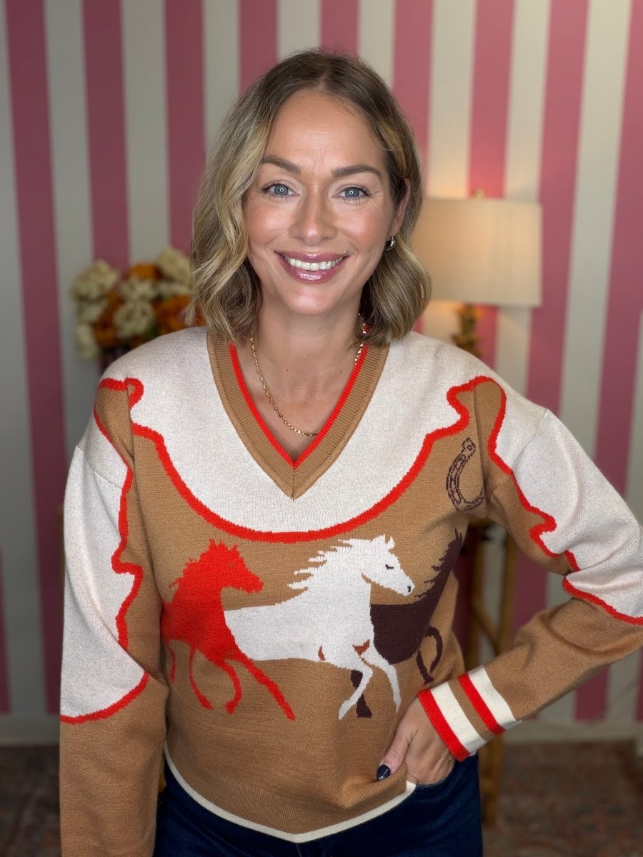 A woman wearing a brown and cream knit sweater with red and white accents, featuring a graphic of a white horse and a red horse on the front, posed in front of a pink and white striped wall.