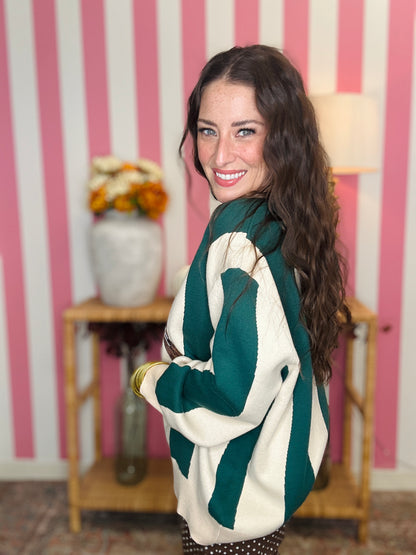 A woman wearing a green and cream striped sweater with a colorful pheasant bird graphic on the front, standing in front of a pink and white striped wall.