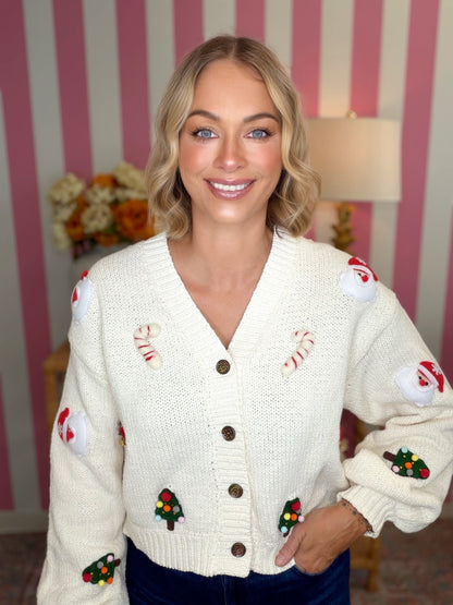 A smiling blonde woman wearing a cream knit holiday cardigan with embroidered candy canes, Santa claus, and Christmas trees, posed in front of pink and white striped wallpaper.