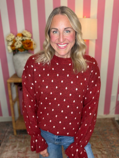 A smiling blonde woman wearing a red knit sweater with a white geometric dot pattern, paired with blue jeans, standing in front of a pink and white striped wall.