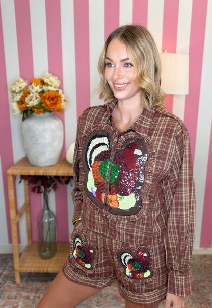 A woman wearing a brown plaid long-sleeve shirt and matching shorts set, both decorated with large sequined turkeys on the front, posed in front of a pink and white striped wall.