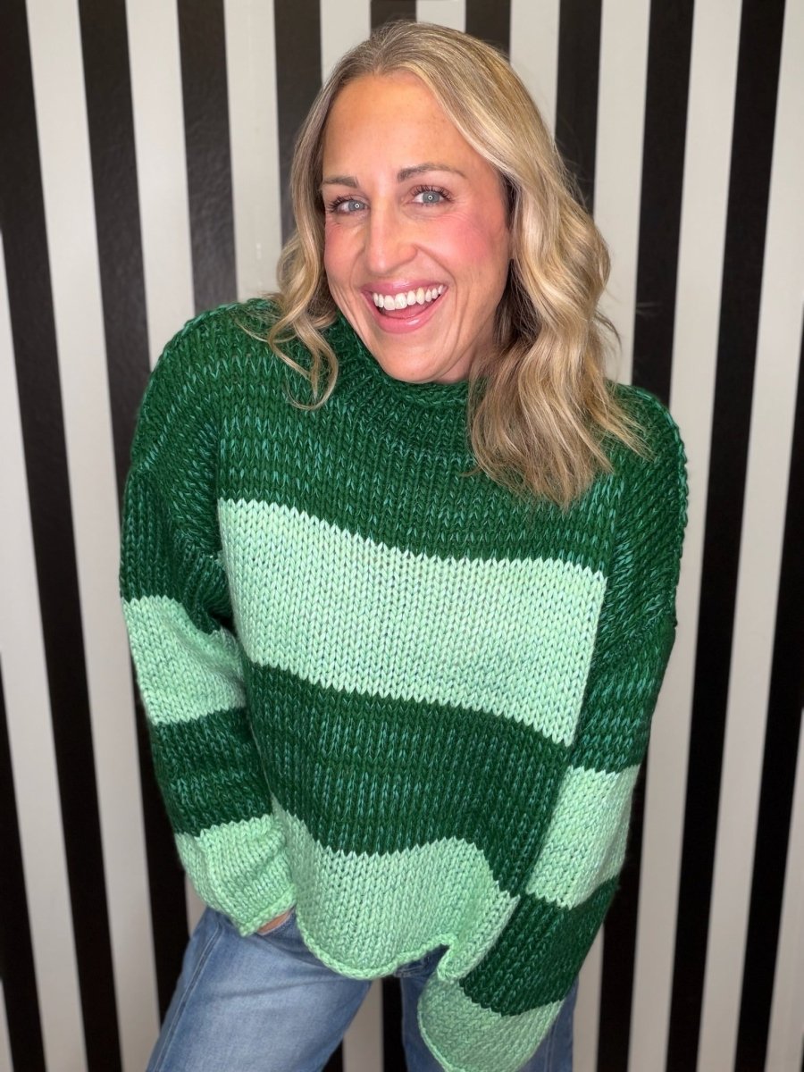 A smiling blonde woman wearing a chunky knit sweater with wide horizontal stripes in dark green and light mint green, paired with blue jeans, standing in front of a black and white vertically striped wall.