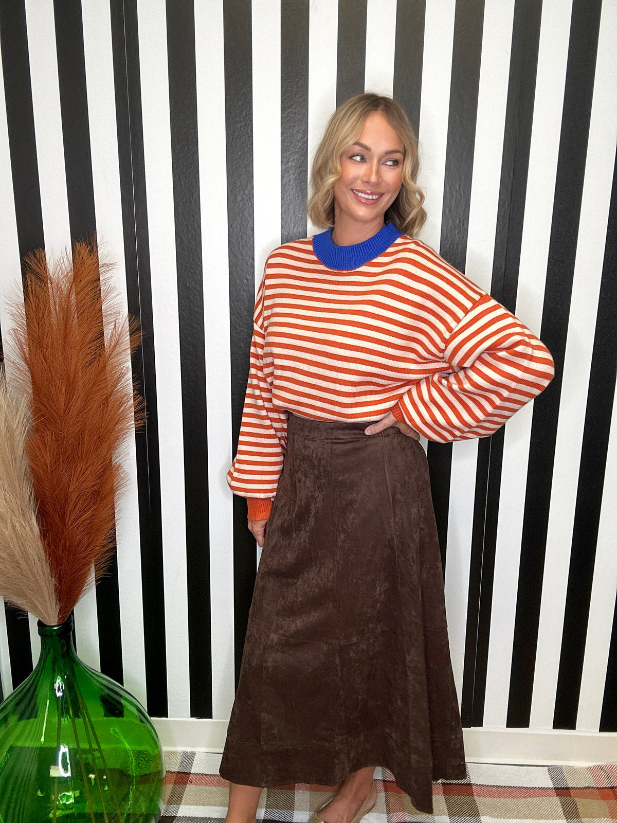 A woman posing in front of a black and white striped wall, wearing a rust-orange and white striped long-sleeve top with a bright blue collar, paired with a brown midi skirt.