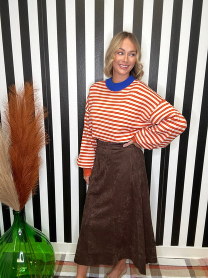 A woman posing in front of a black and white striped wall, wearing a rust-orange and white striped long-sleeve top with a bright blue collar, paired with a brown midi skirt.