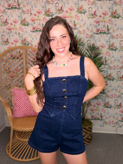 Woman wearing a denim romper standing in front of a patterned wall.
