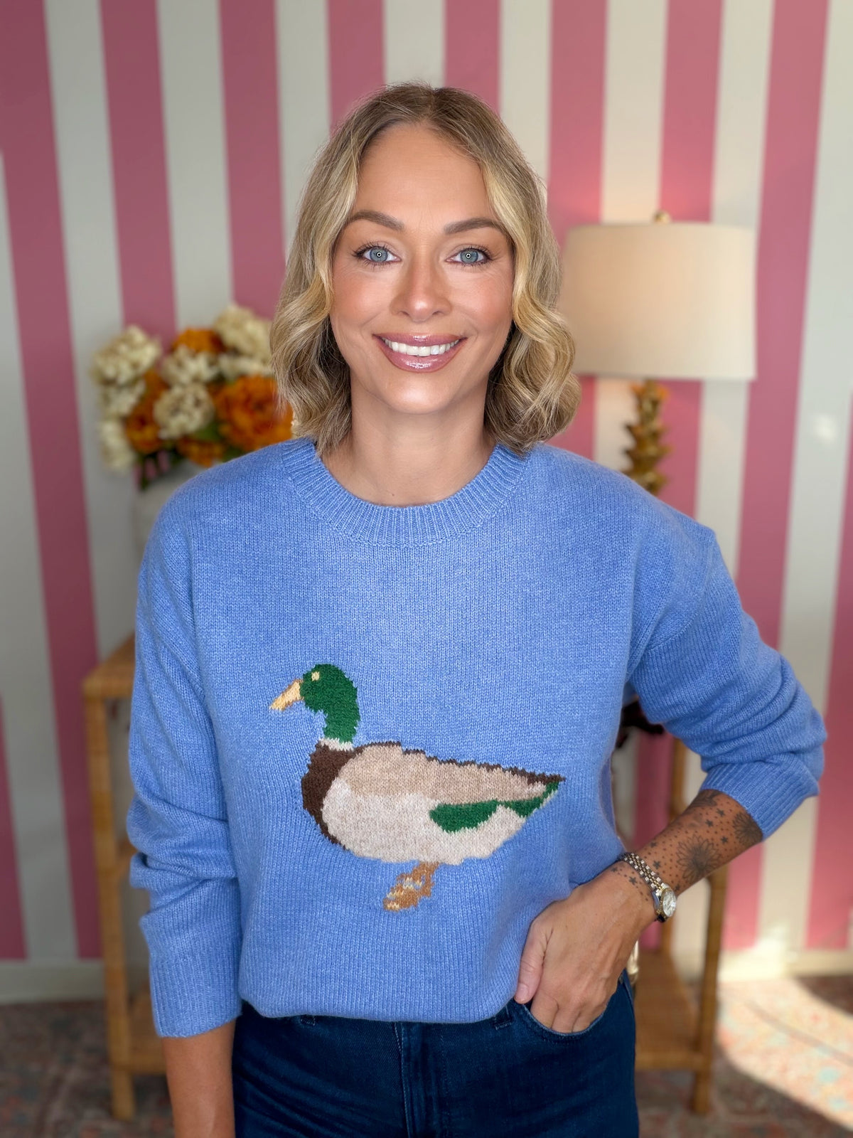 A smiling blonde woman wearing a light blue crewneck sweater with a large knit mallard duck design on the front, paired with dark blue jeans, standing in front of a pink and white striped wall.