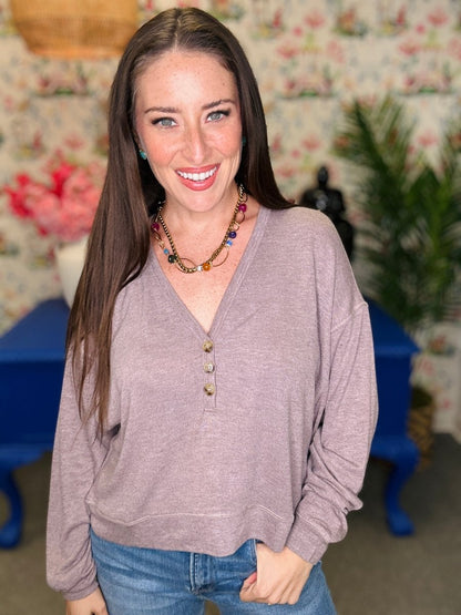 A smiling woman with long brown hair wearing a heathered mauve-purple long-sleeve henley top and blue jeans, standing in a decorated room with floral wallpaper, a blue table, and green plants.