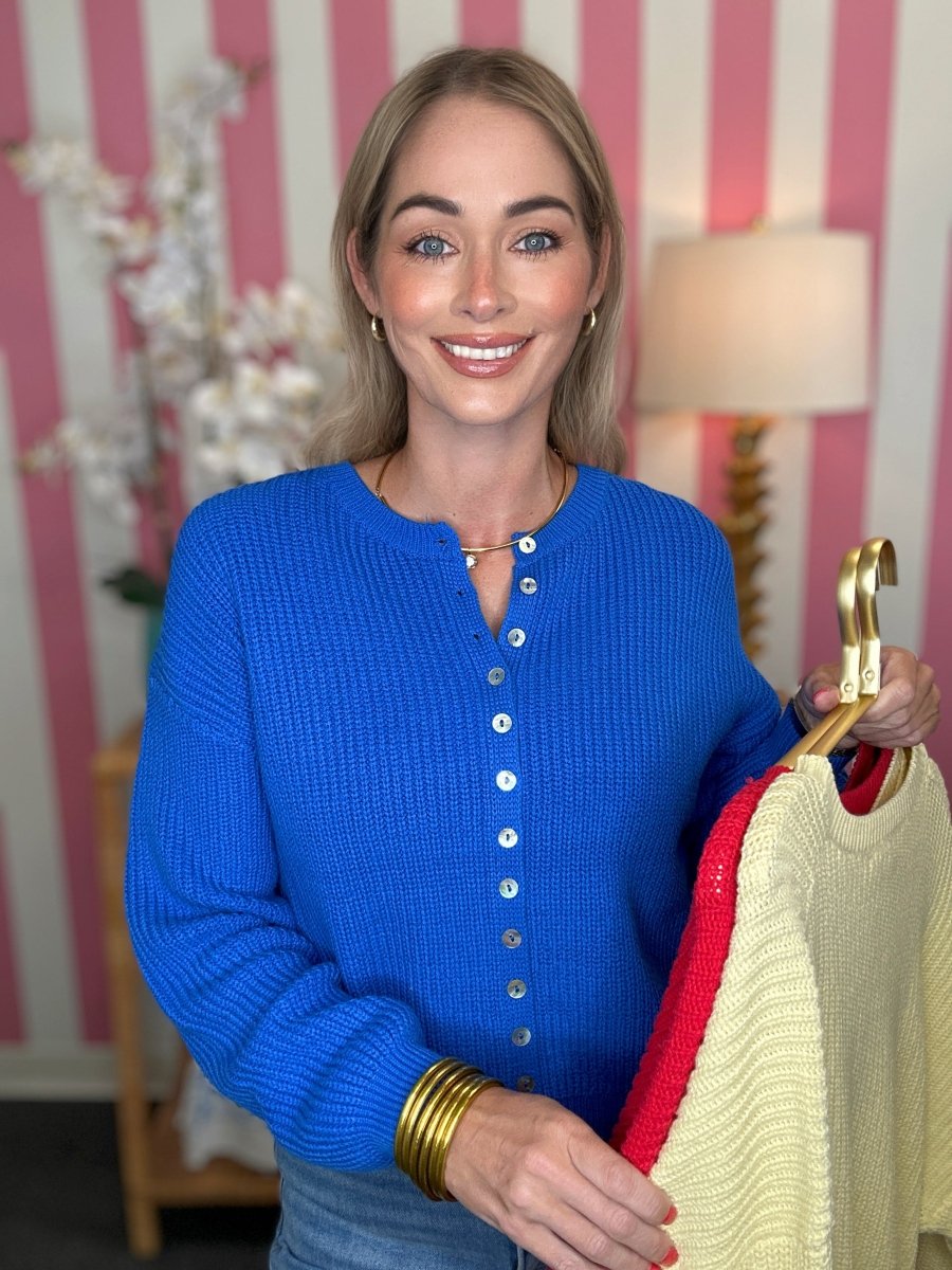 A smiling woman wearing a bright blue ribbed knit cardigan sweater holds up a cream knit sweater on a gold hanger with a red trim, set against a pink and white striped wall background.