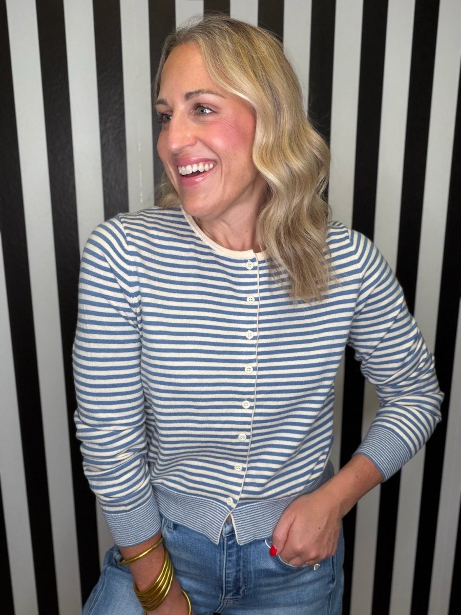 A woman with blonde hair smiling, wearing a blue and white striped long-sleeve button-up cardigan and blue jeans, standing in front of a black and white striped wall.