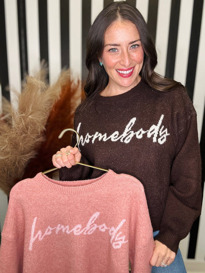 A woman holds up a dusty pink knit sweater with 'homebody' written across the chest on a hanger, while wearing a dark brown sweater with the same script behind her. The background features a black and white striped wall with dried pampas grass on the left.