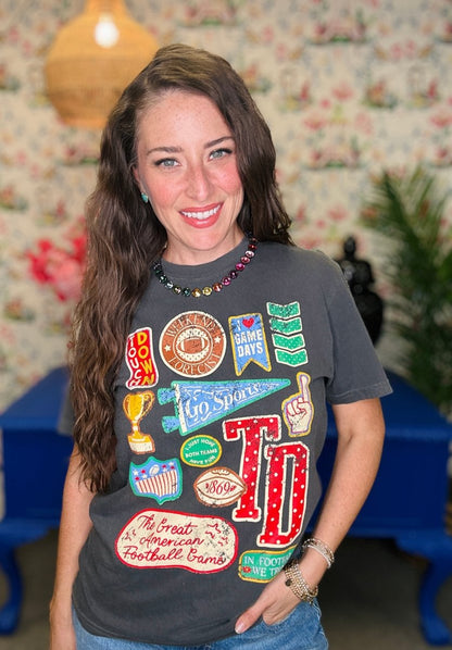 A smiling woman with long brown hair wearing a dark gray graphic t-shirt with colorful sports-themed patches and 'TD' lettering, paired with blue jeans in a decorated indoor setting.