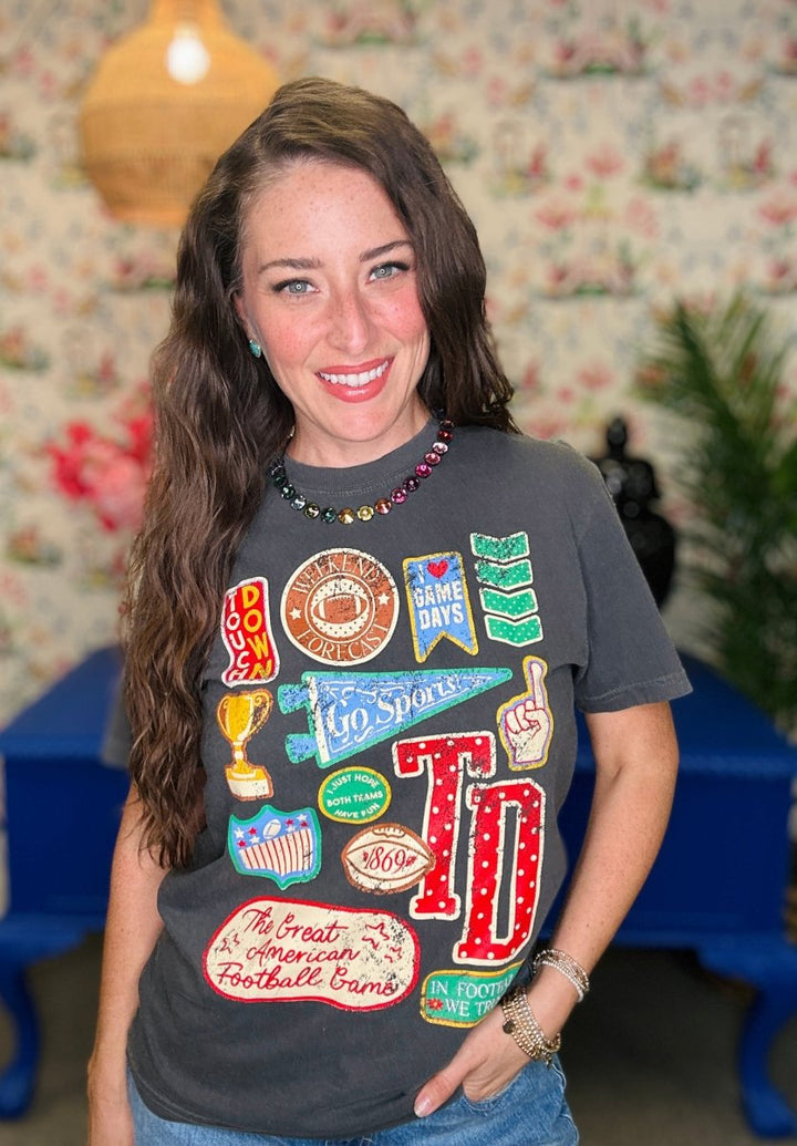 A smiling woman with long brown hair wearing a dark gray graphic t-shirt with colorful sports-themed patches and 'TD' lettering, paired with blue jeans in a decorated indoor setting.