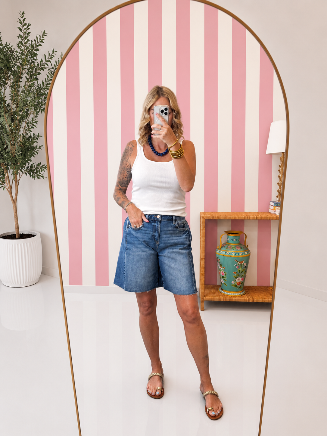 Person taking a mirror selfie wearing a white tank top and blue shorts in a room with pink and white striped wallpaper.