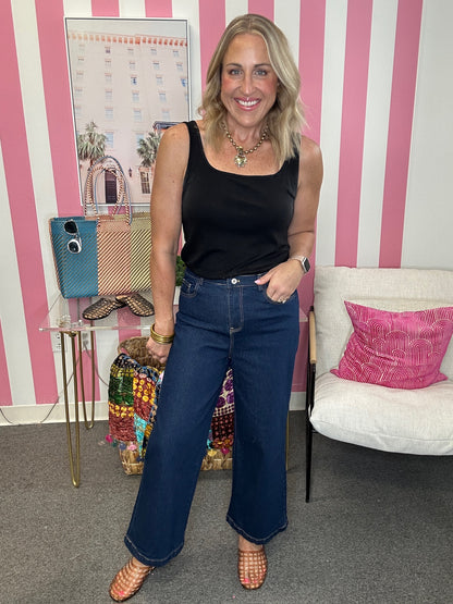 A woman posing in a black tank top and dark blue wide-leg jeans, standing in a room with pink and white vertical stripes on the wall.