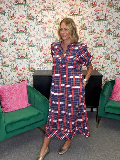 A woman modeling a navy blue midi dress with a red and white plaid pattern, standing in a room with floral wallpaper, green velvet armchairs, and a black cabinet.