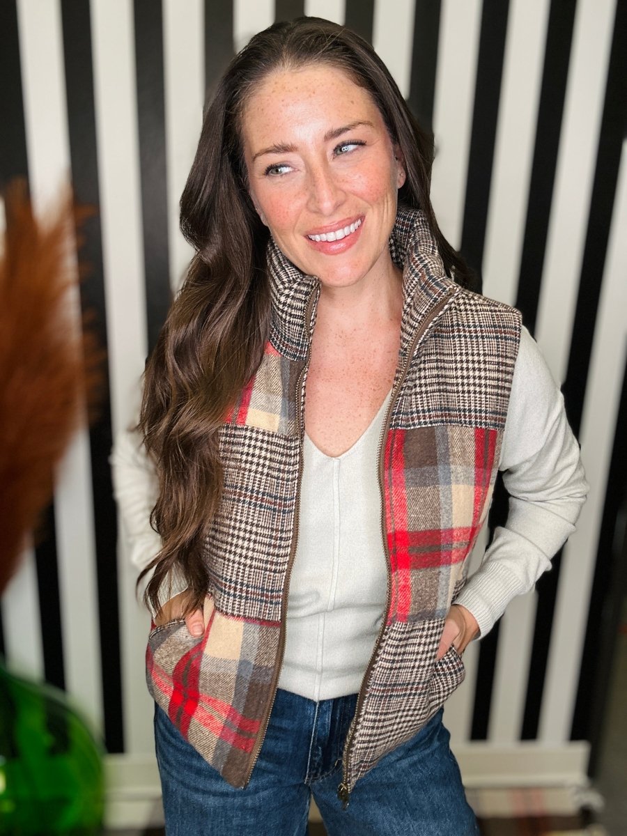A woman wearing a sleeveless plaid vest over a white top and blue jeans, standing in front of a black and white striped background.